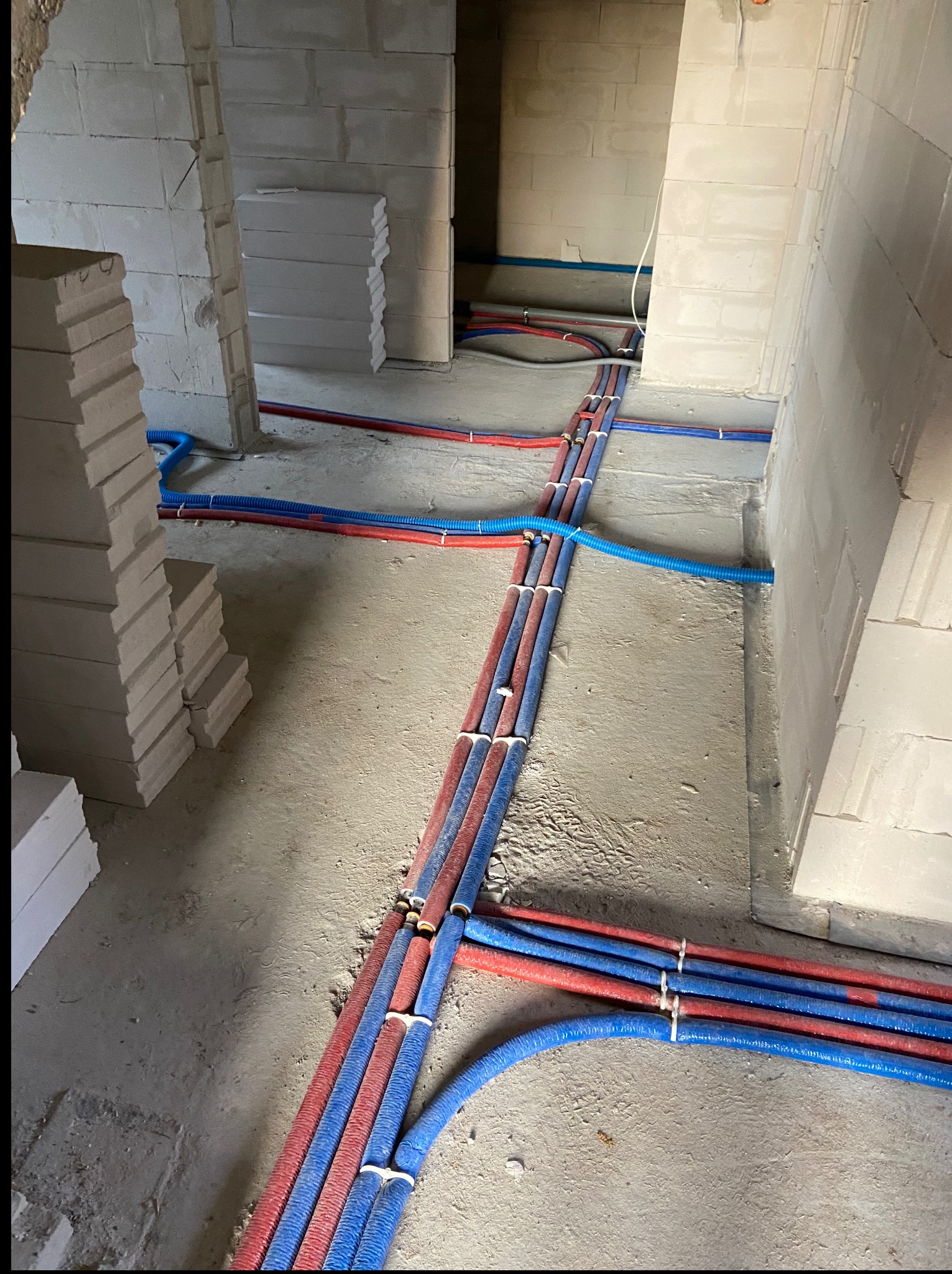 Nowa instalacja wodno-kanalizacyjna i CO w surowym budynku. Czerwone i niebieskie rury prowadzone po podłodze, przymocowane opaskami. Widoczne białe bloczki.