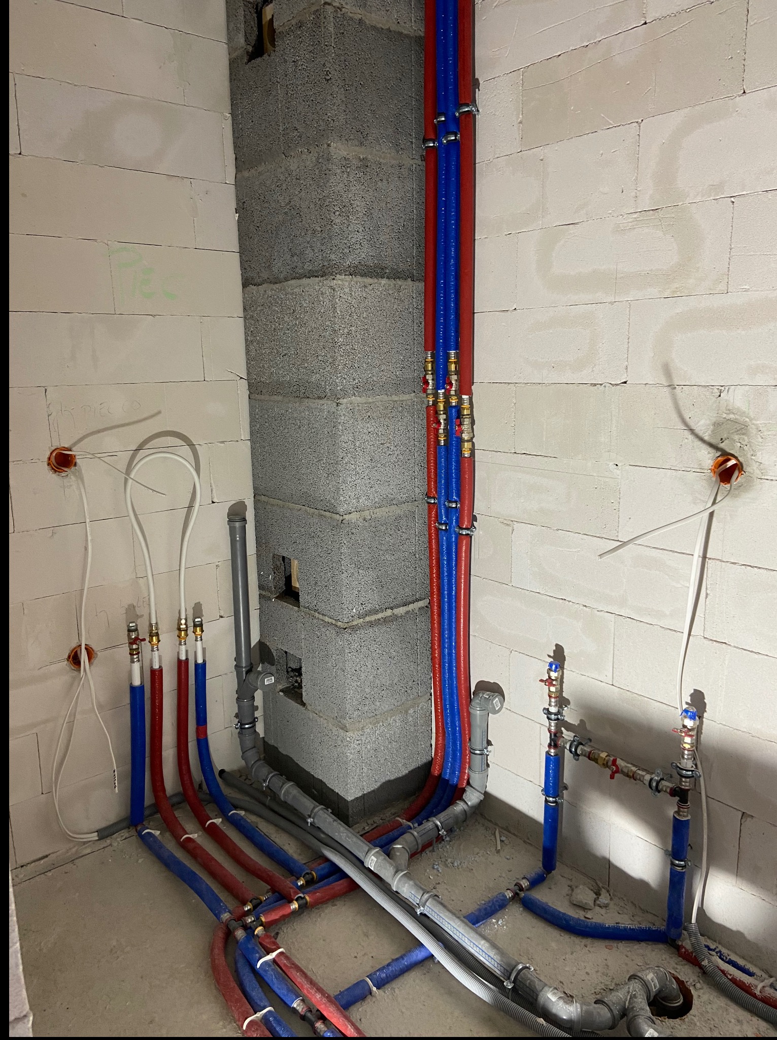 Instalacja wodna z rur PEX w kolorach niebieskim i czerwonym, z mosiężnymi złączkami, zamocowana do ściany z betonu komórkowego i szarego komina wentylacyjnego.