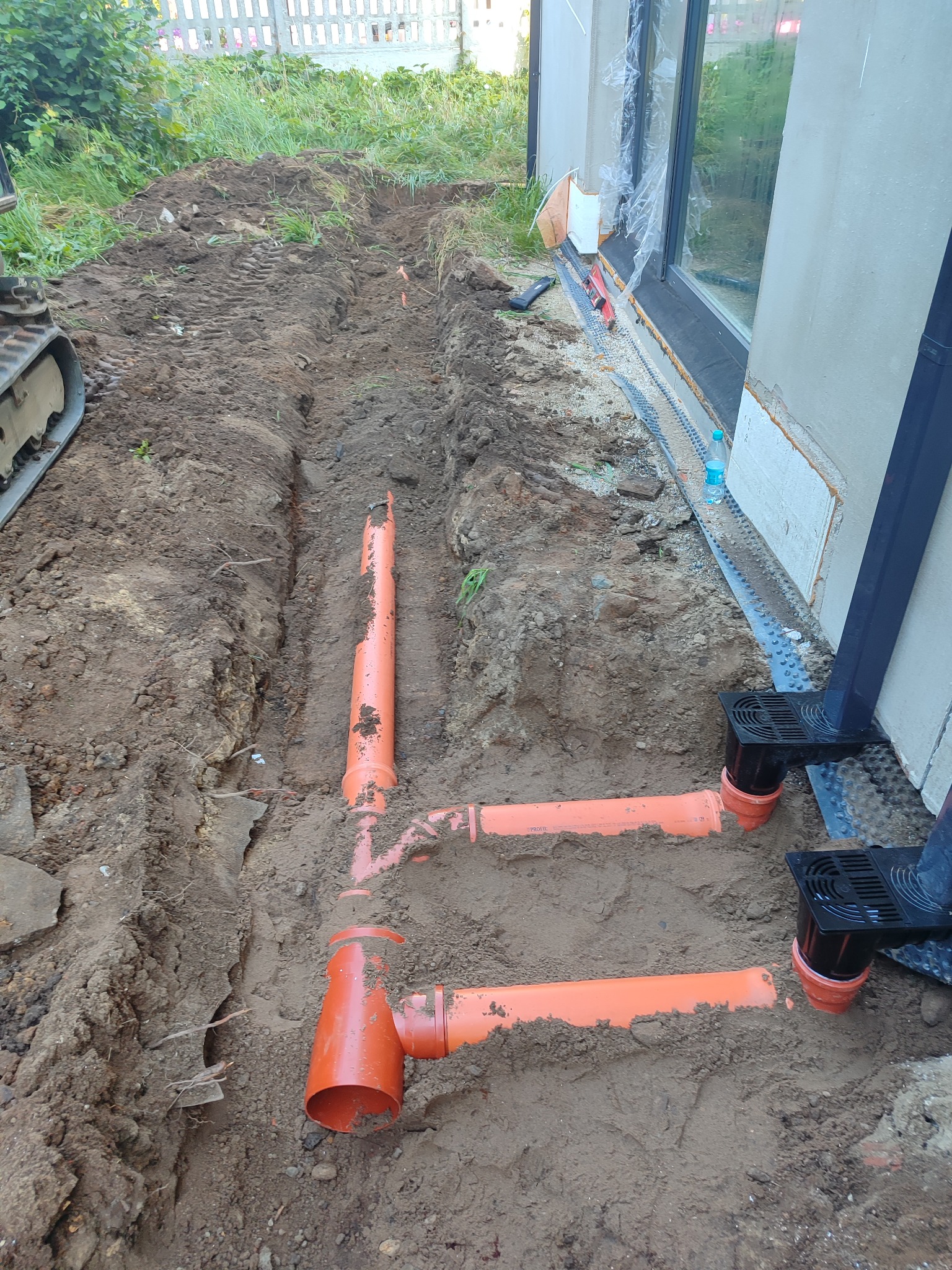 Pomarańczowe rury kanalizacyjne ułożone w wykopie ziemnym wzdłuż fundamentów budynku, widoczne studzienki odpływowe i częściowo odsłonięty fundament.