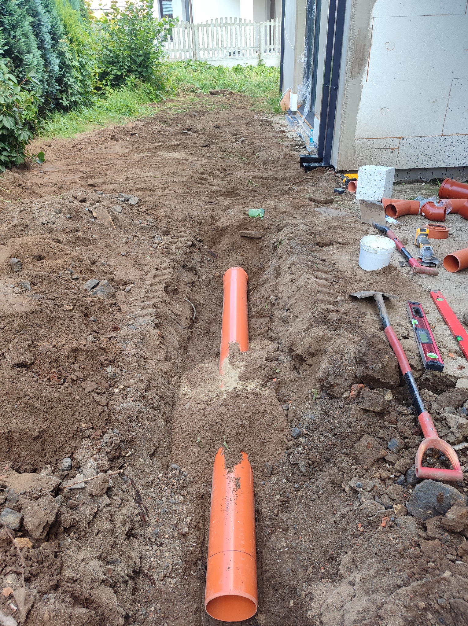 Wykopany rów pod pomarańczowe rury instalacyjne, widoczne narzędzia: szpadel, poziomica, kielnia, obok budynek w trakcie budowy.
