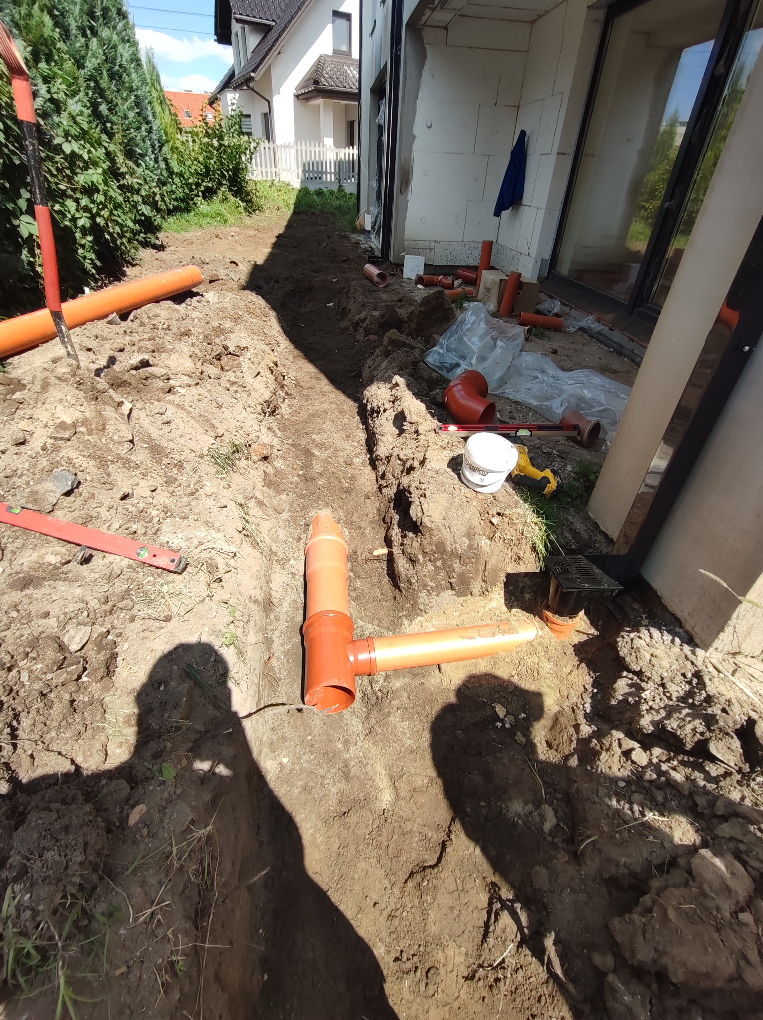 Wykop w ziemi z widocznymi pomarańczowymi rurami kanalizacyjnymi, poziomica, wiadro z klejem i narzędzia w tle, cień osoby w kadrze.