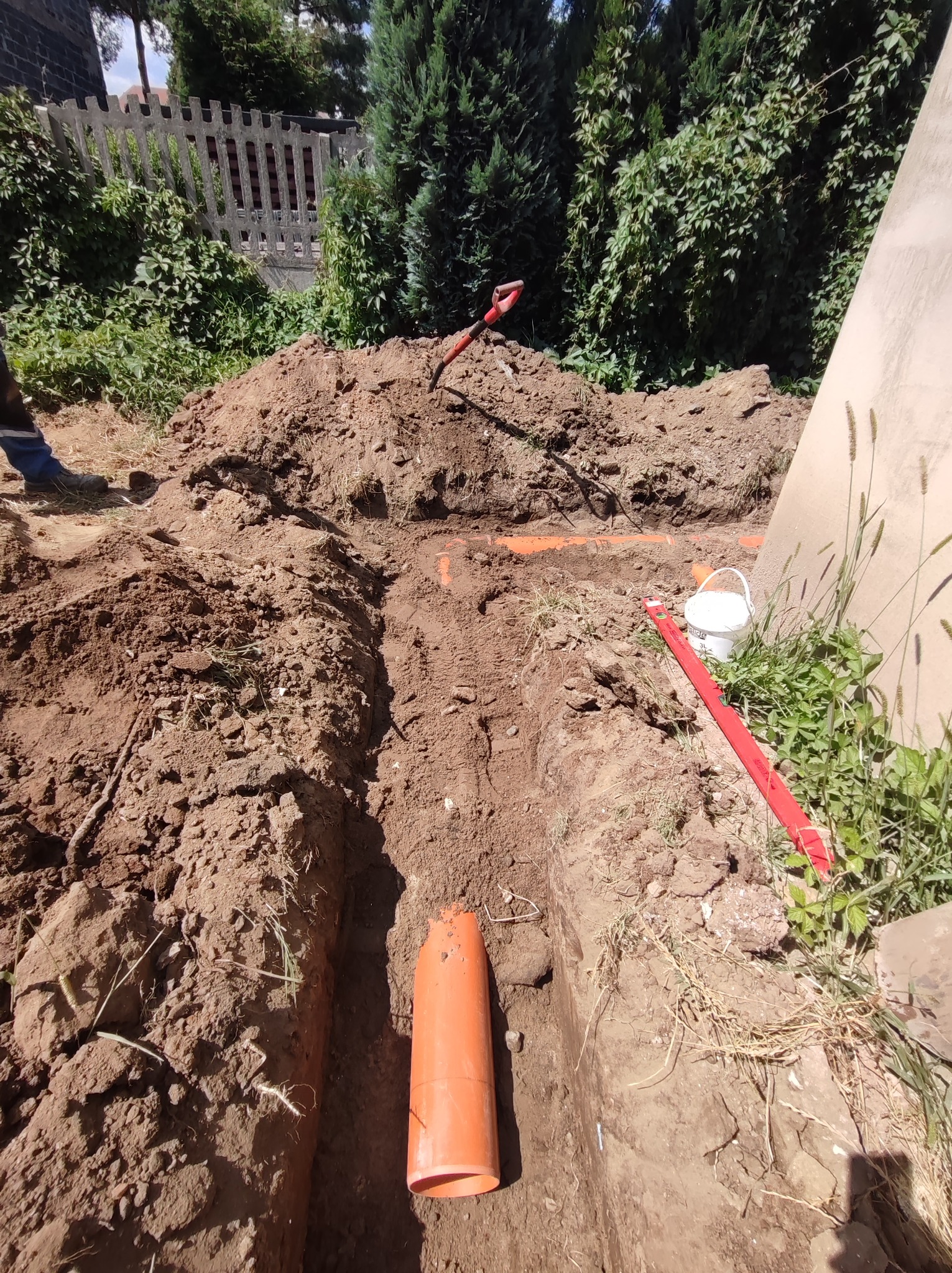 Wykopany rów z pomarańczowymi rurami instalacyjnymi w ziemi, widoczna łopata, poziomica i wiadro, w tle zielona roślinność i drewniane ogrodzenie.