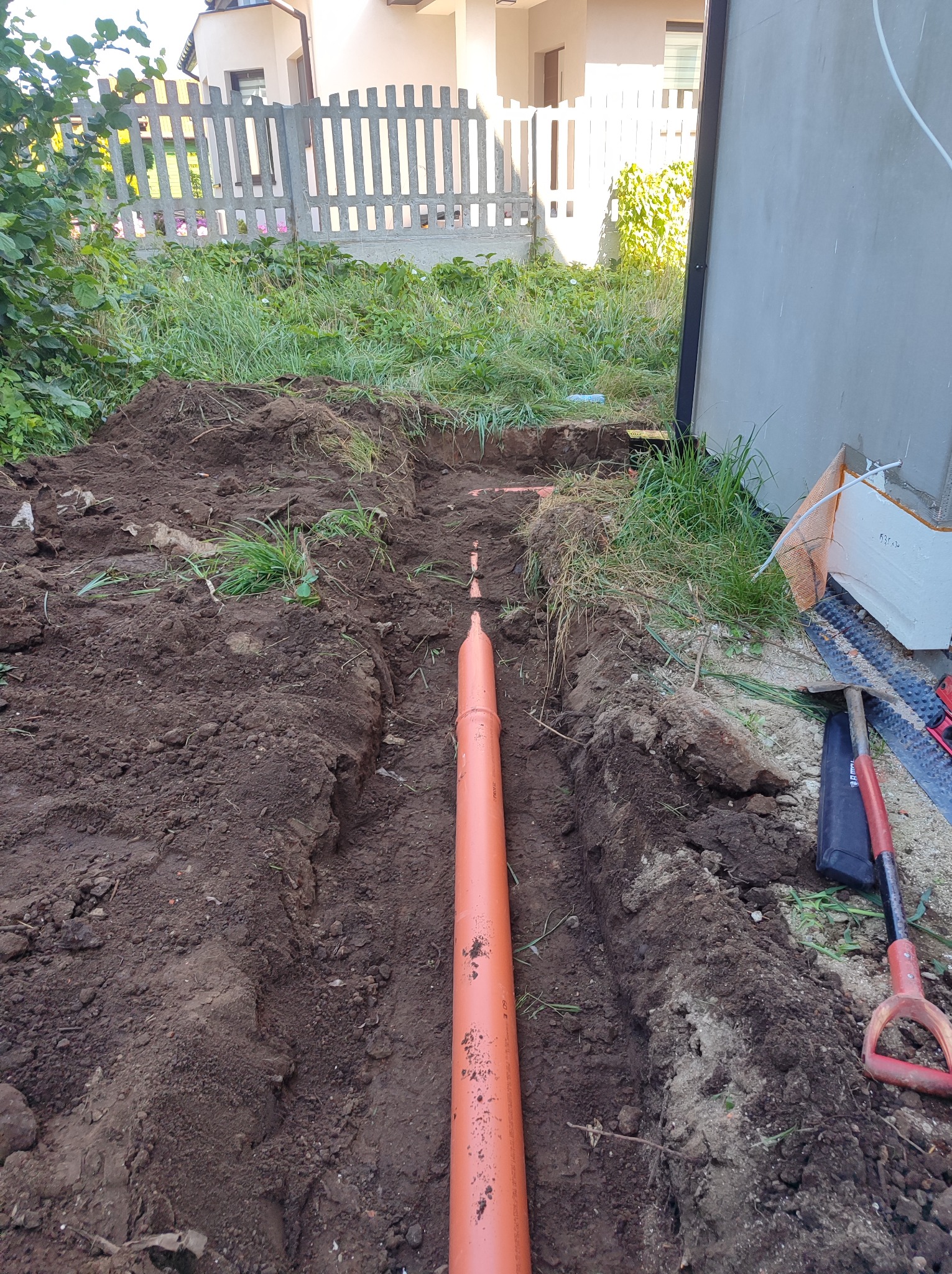 Pomarańczowa rura ułożona w wykopie ziemnym, widoczne narzędzia i częściowo budynek w tle.