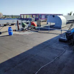 Pracownik w pomarańczowej koszulce na płaskim dachu, przy pracy z butlą gazową i palnikiem, wentylacja w tle, widok na miasto.