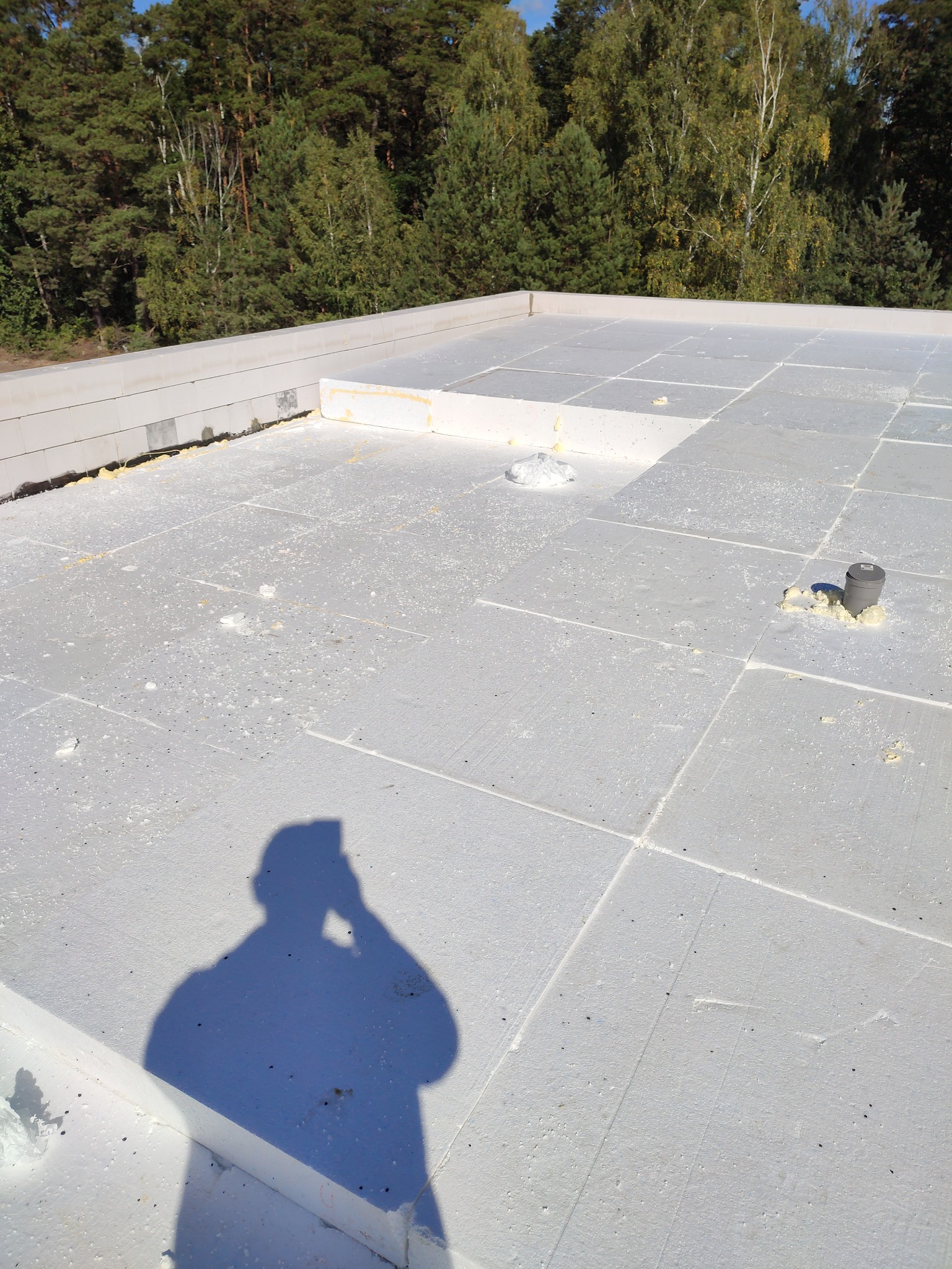 Płaski dach pokryty szarymi płytami izolacyjnymi, widoczne łączenia i wystający kominek wentylacyjny, w tle las, cień osoby robiącej zdjęcie.