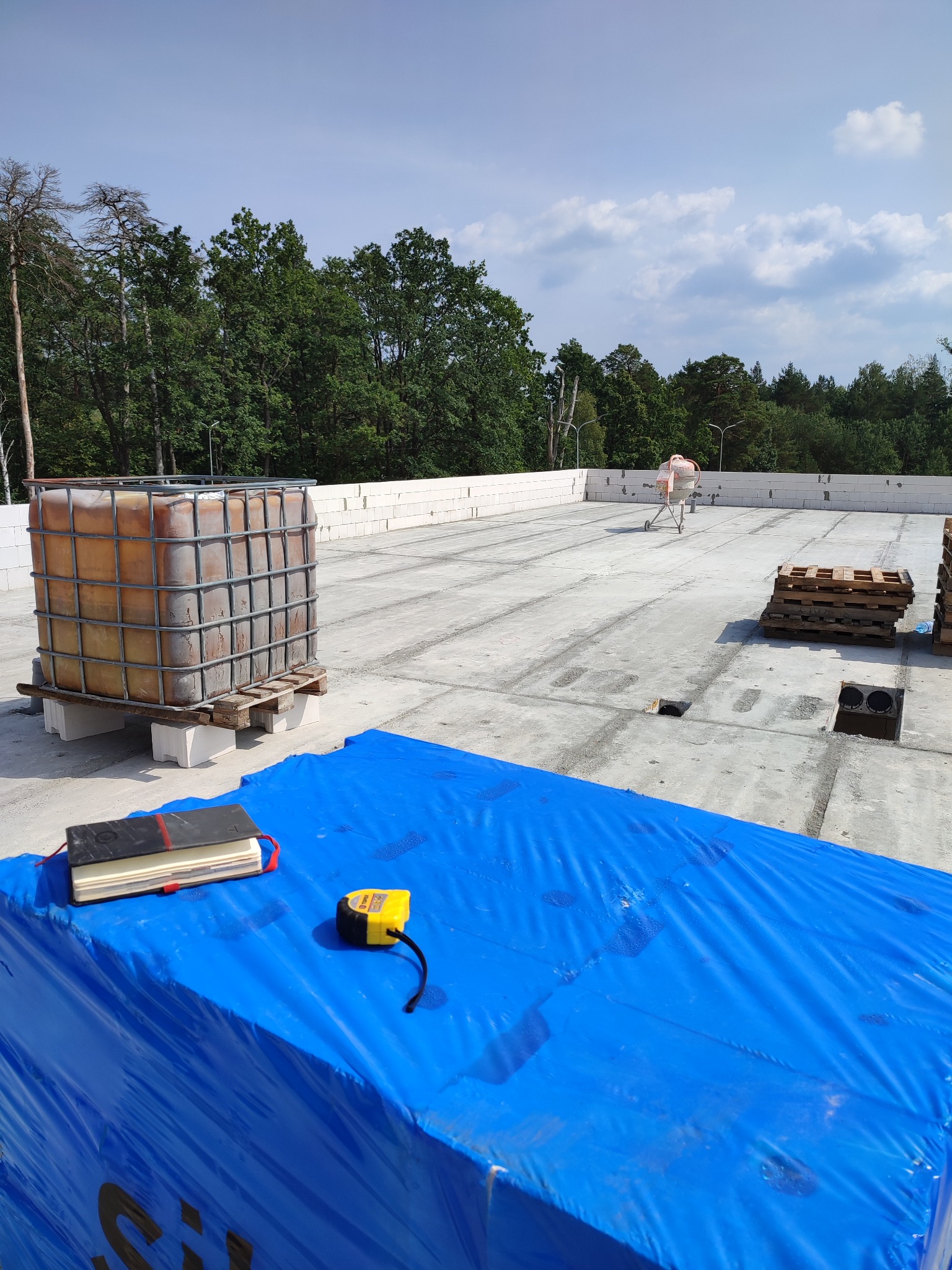 Widok z góry na płaski dach budynku w trakcie budowy, z widocznym zbiornikiem na wodę w metalowej klatce, paletami, betonową mieszarką w tle, zwiniętą folią w kolorze niebieskim, notesem i miarką...
