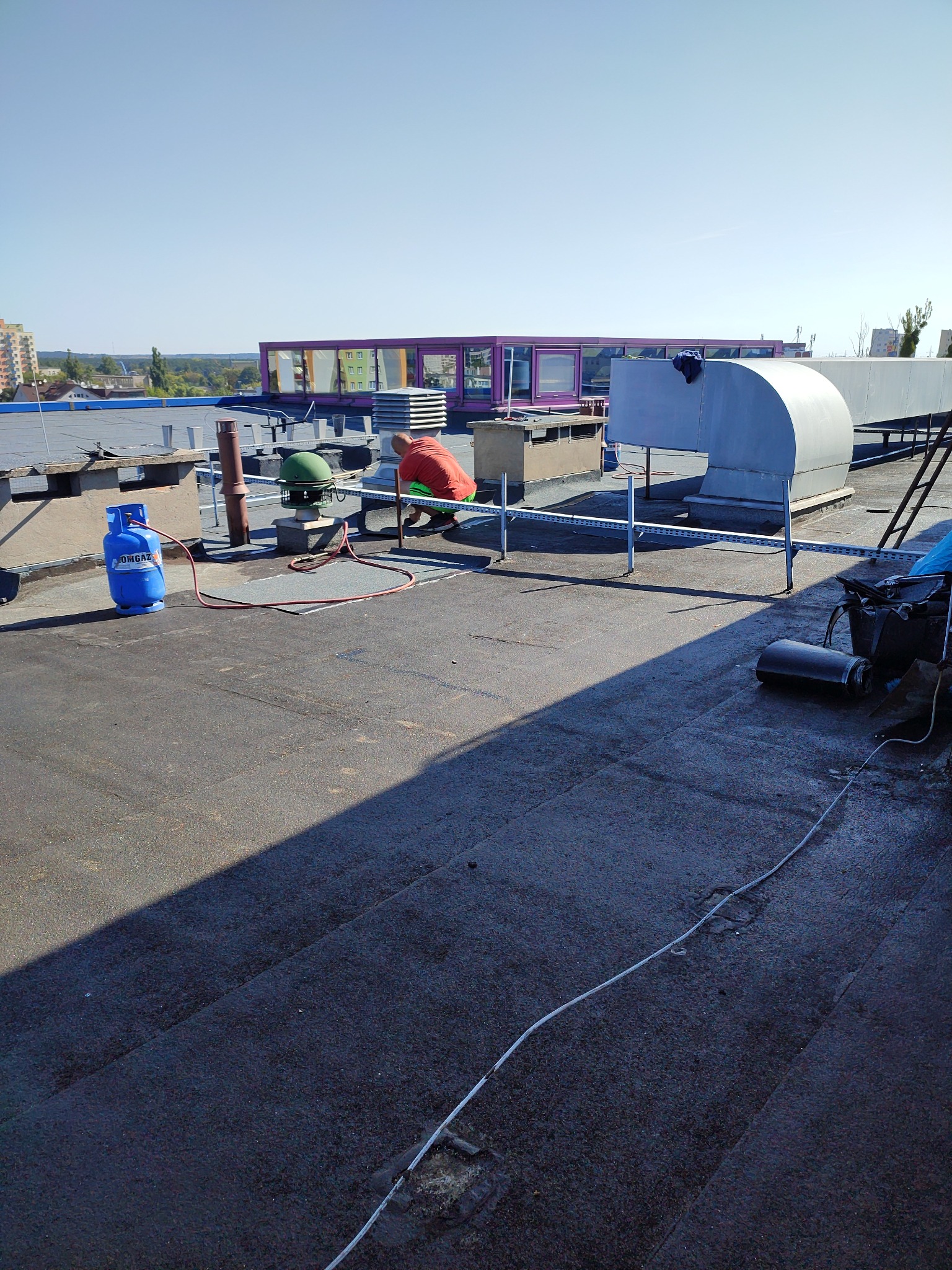 Pracownik w pomarańczowej koszulce na płaskim dachu, przy pracy z butlą gazową i palnikiem, wentylacja w tle, widok na miasto.