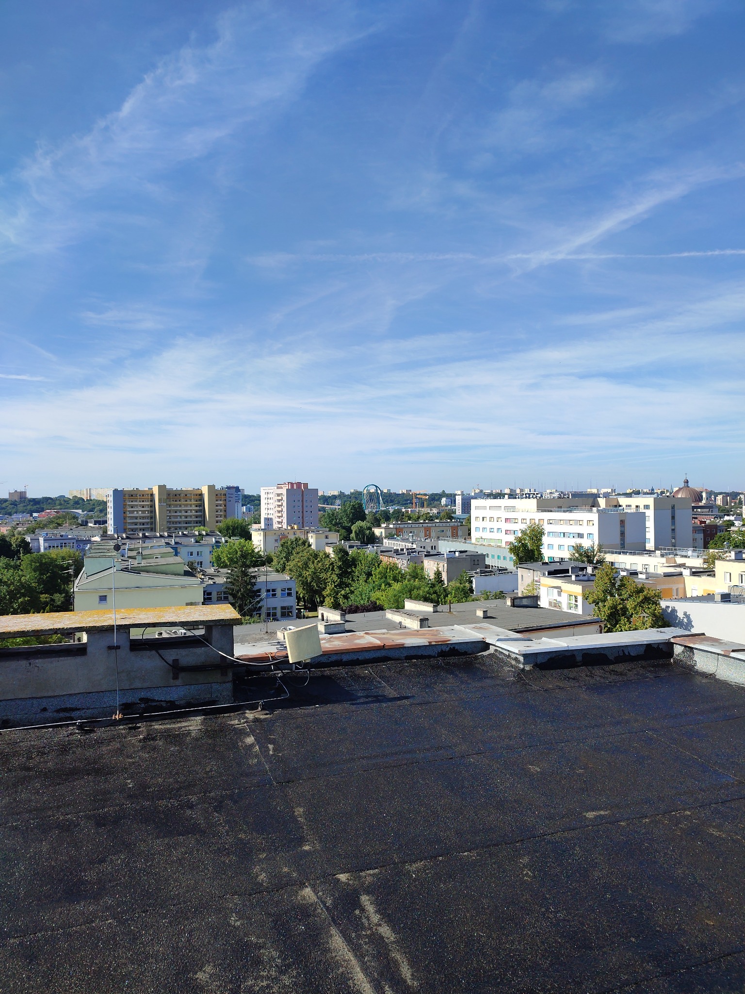 Widok z dachu pokrytego czarną papą na panoramę miasta w słoneczny dzień, widoczne budynki mieszkalne i drzewa.