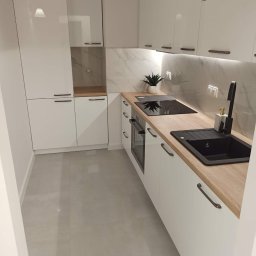 Przemysław Czuchra - Nowoczesna, biała kuchnia z drewnianym blatem, czarnym zlewem i płytą indukcyjną, marmurowym panelem ściennym oraz szarymi płytkami podłogowymi.