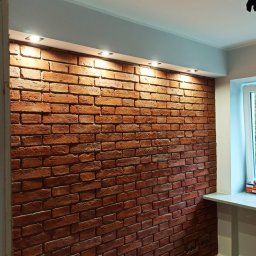 Przemysław Czuchra - Ściana z czerwonej cegły dekoracyjnej oświetlona punktowymi lampami sufitowymi, widoczny fragment okna i podłogi z paneli.