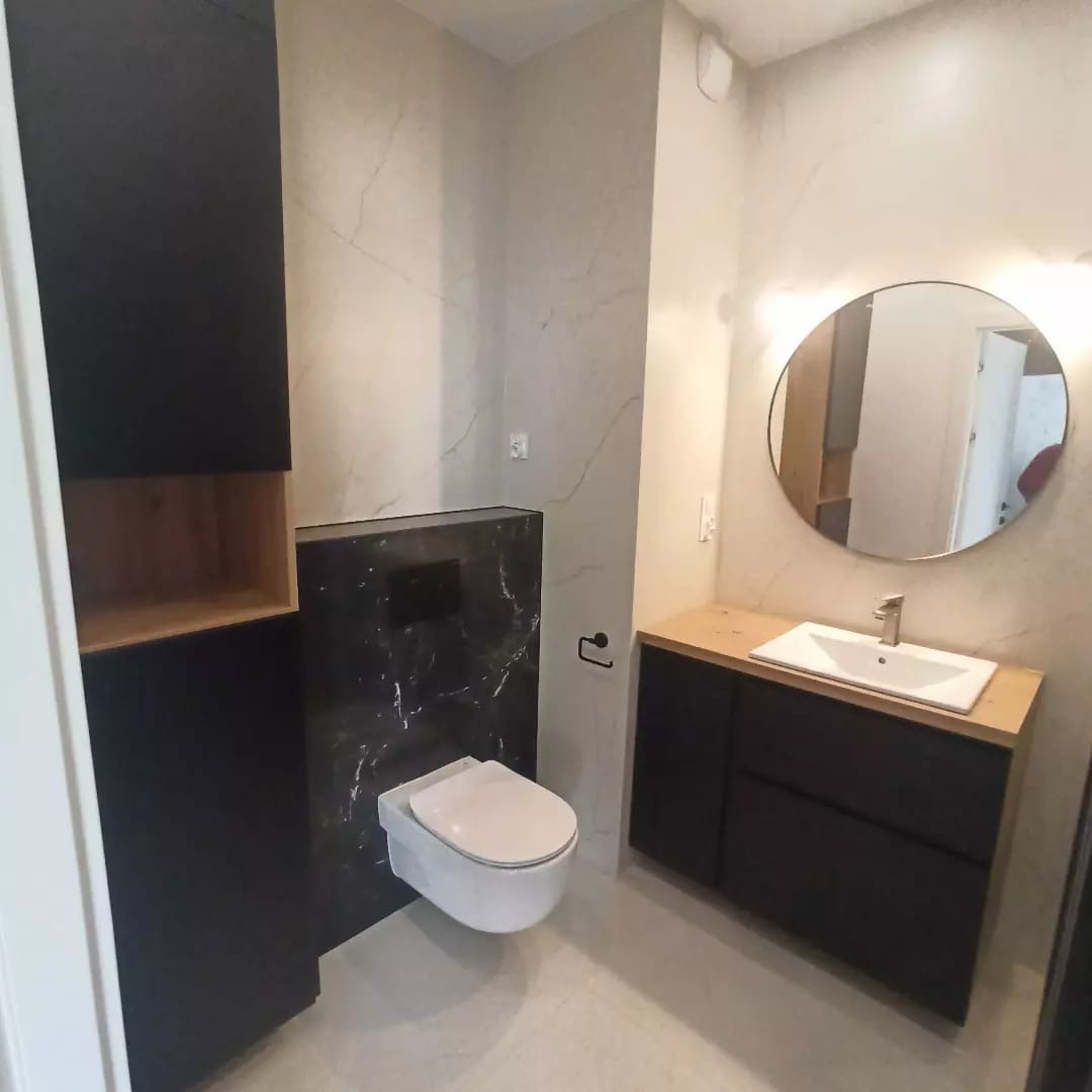 Nowoczesna łazienka z marmurowymi płytkami, wiszącą toaletą na czarnym tle i okrągłym lustrem z podświetleniem nad umywalką z szafką w kolorze czarnym i blatem w kolorze drewna.