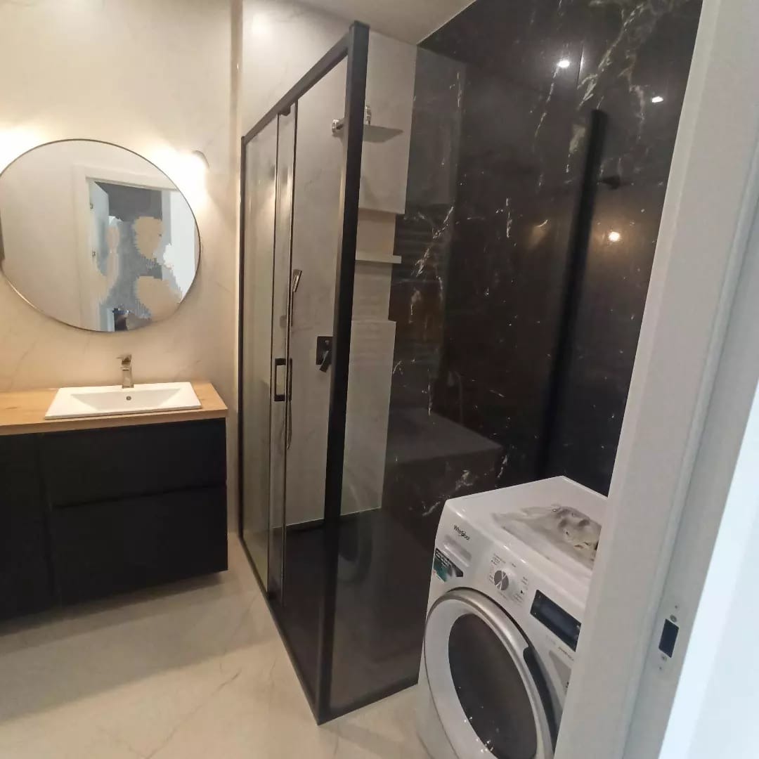 Nowoczesna łazienka z kabiną prysznicową typu walk-in z czarnymi ramami, umywalką na szafce z drewnianym blatem, okrągłym lustrem podświetlanym od tyłu i pralką Whirlpool.