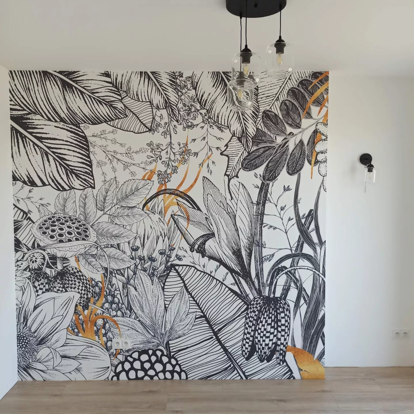 Ściana z fototapetą w czarno-białe tropikalne liście z elementami złota, oświetlona trzema wiszącymi lampami z przezroczystymi kloszami i czarnymi oprawkami, na podłodze panele w odcieniach beżu.