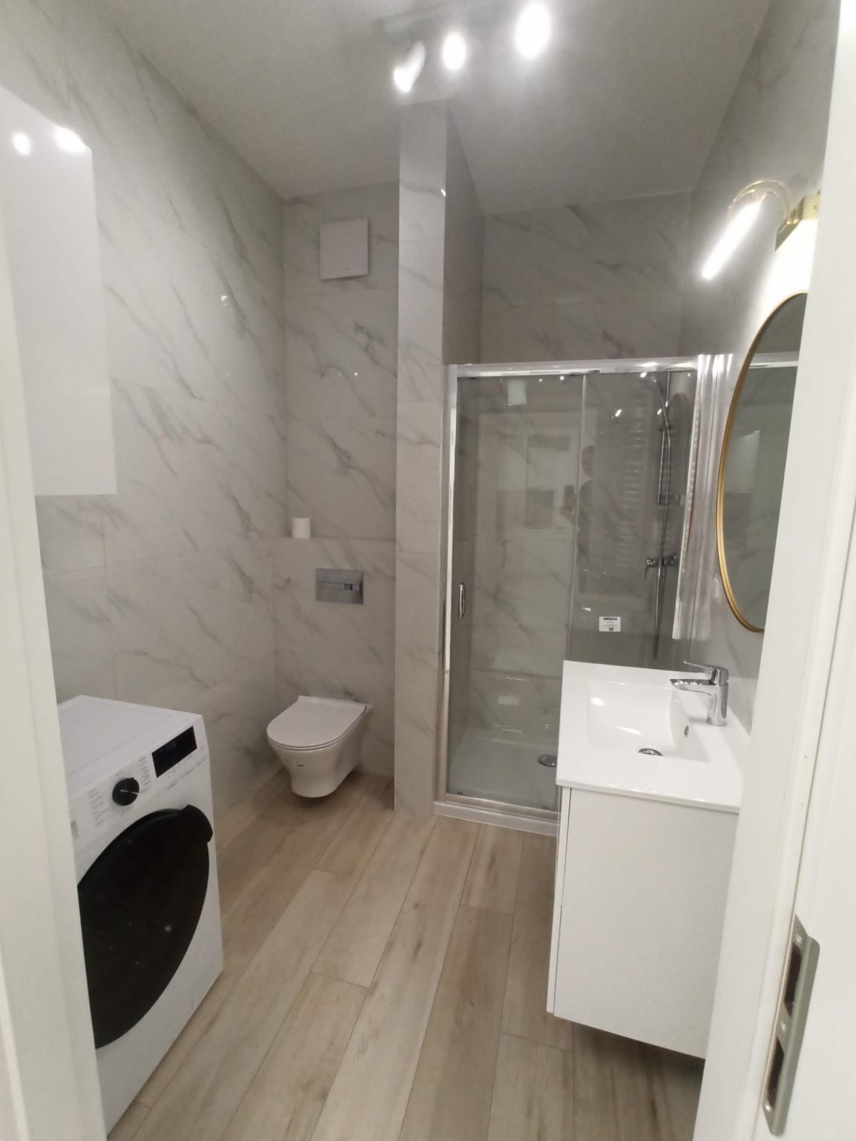 Nowoczesna łazienka w odcieniach bieli i szarości z marmurowymi płytkami na ścianach, drewnopodobną podłogą, kabiną prysznicową ze szklanymi drzwiami, umywalką z szafką, toaletą podwieszaną i pralką.