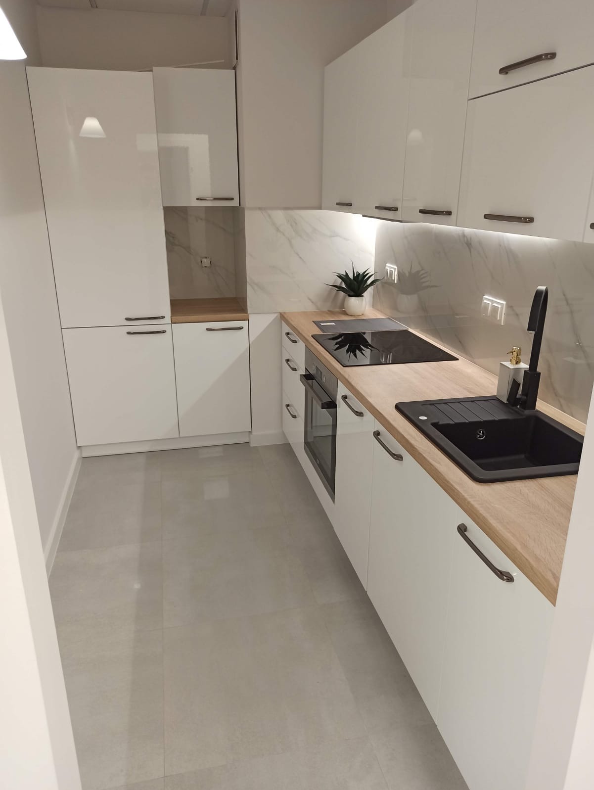 Nowoczesna, biała kuchnia z drewnianym blatem, czarnym zlewem i płytą indukcyjną, marmurowym panelem ściennym oraz szarymi płytkami podłogowymi.