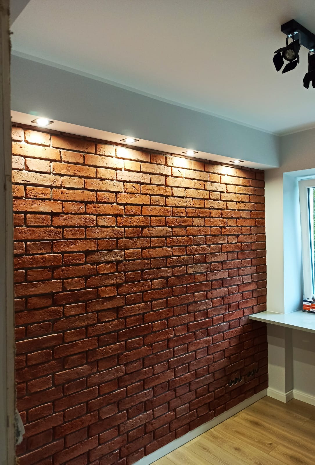 Ściana z czerwonej cegły dekoracyjnej oświetlona punktowymi lampami sufitowymi, widoczny fragment okna i podłogi z paneli.