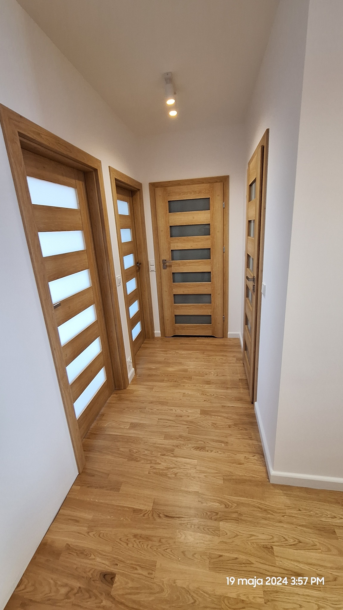 Korytarz z trzema drewnianymi drzwiami z poziomymi przeszkleniami, jasna podłoga drewniana, białe ściany i punktowe oświetlenie sufitowe.