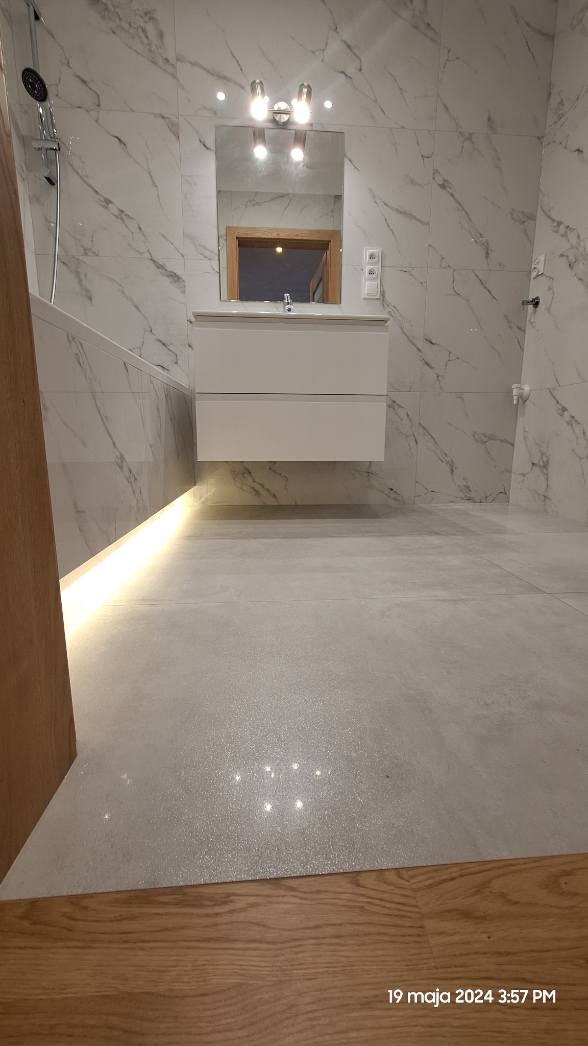Nowoczesna łazienka z białymi marmurkowymi płytkami, podświetlaną wanną, wiszącą szafką z umywalką i lustrem z drewnianą ramą, oświetlona lampami nad lustrem. Widoczne gniazdka elektryczne na ścianie.