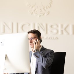 NICIŃSKI Kancelaria Restrukturyzacyjna Tomasz Niciński - Prawnik w okularach rozmawia przez telefon w biurze w Łodzi. W tle godło Polski i nazwa kancelarii na ścianie, widoczny fragment komputera.