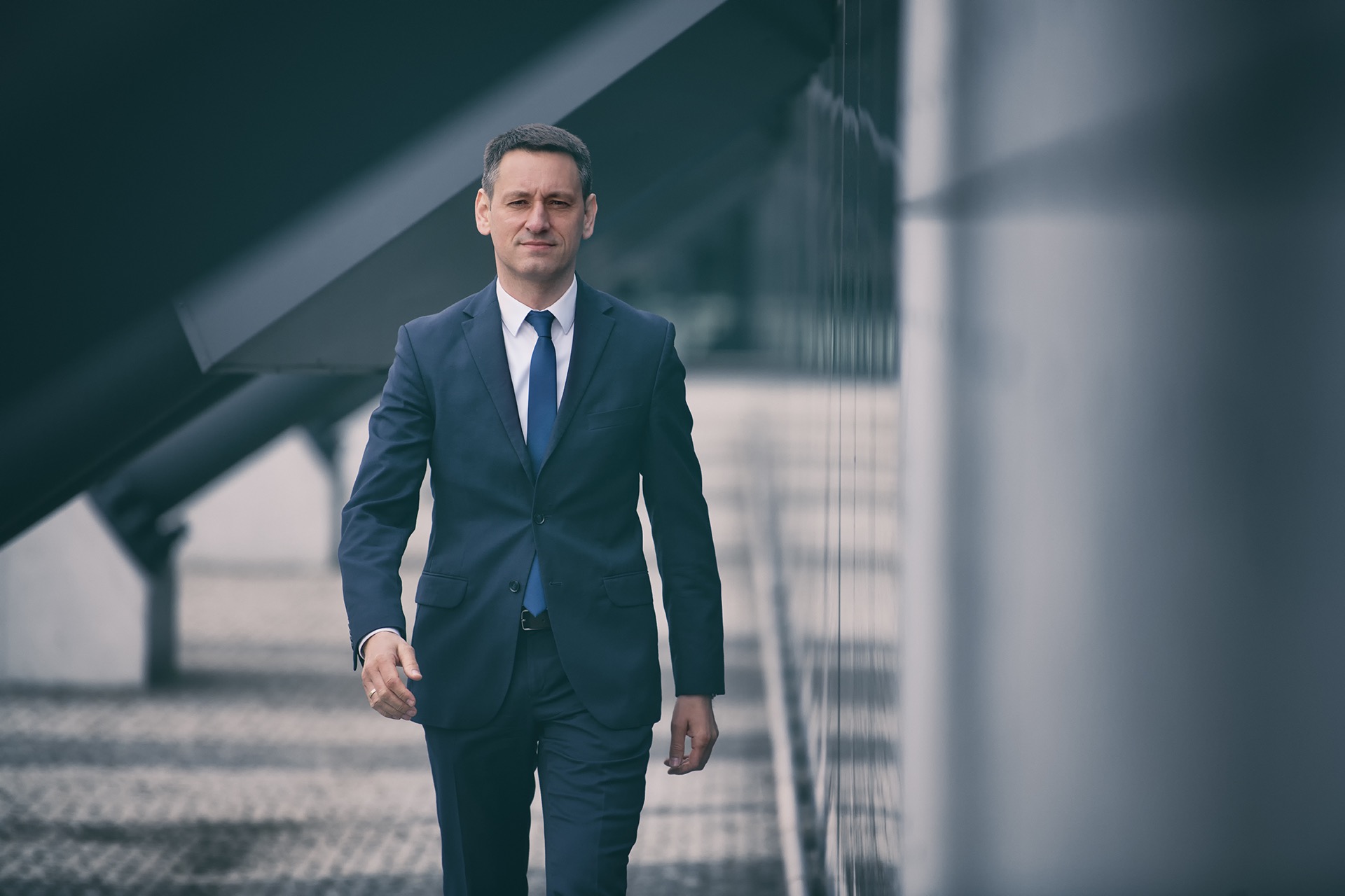 Mężczyzna w granatowym garniturze i niebieskim krawacie idzie w kierunku kamery na tle nowoczesnego budynku z elementami konstrukcyjnymi.