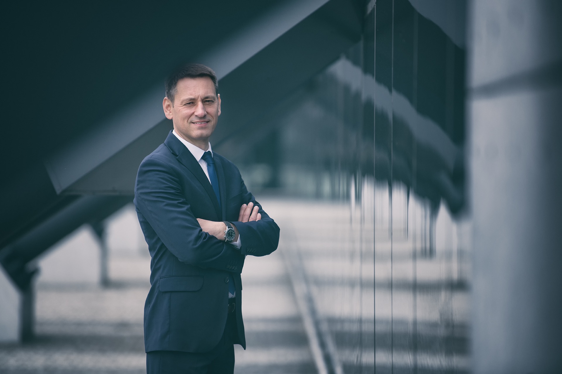 Uśmiechnięty prawnik w granatowym garniturze, z zegarkiem na ręce, stoi na tle nowoczesnej architektury.