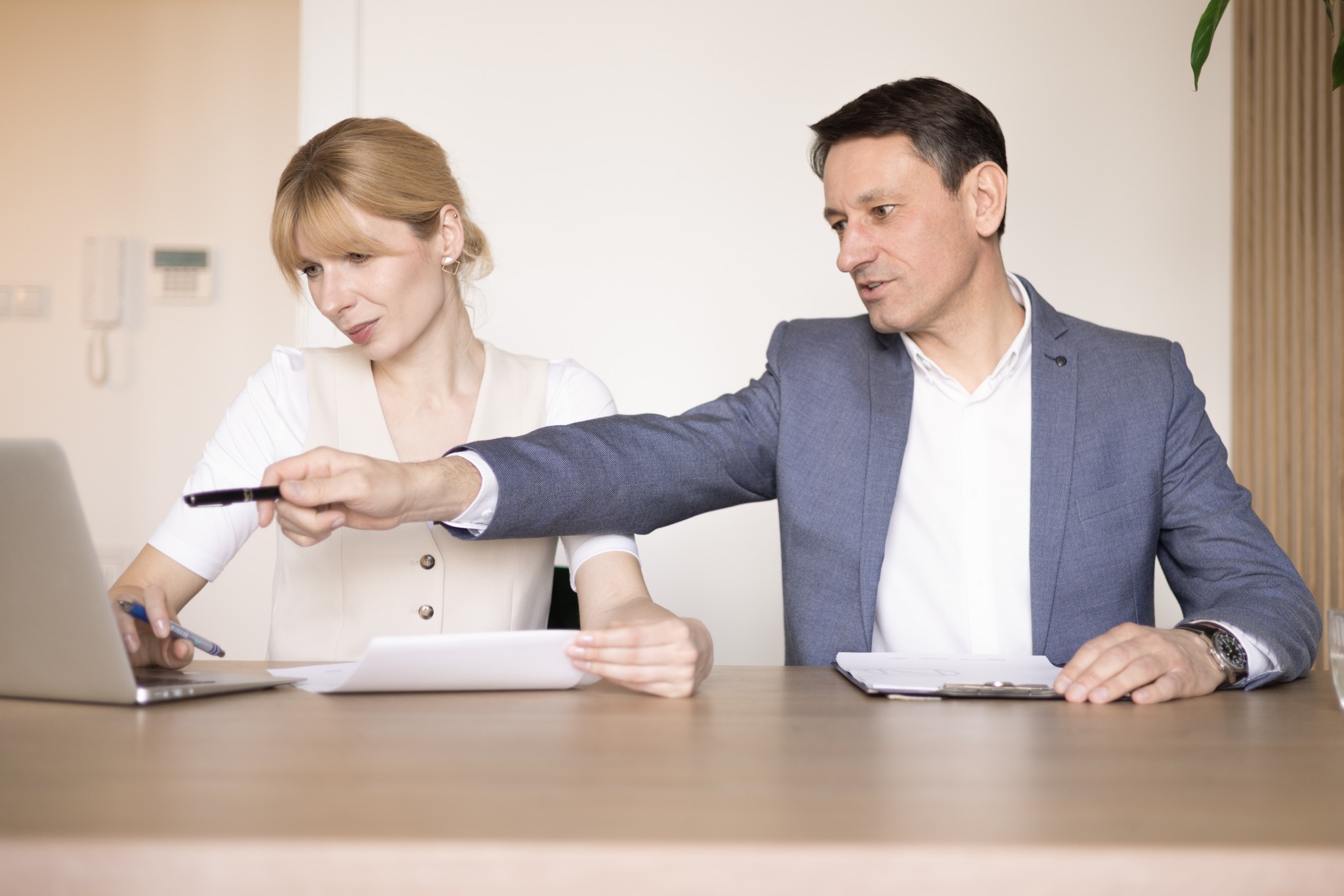 Konsultacja biznesowa: mężczyzna w garniturze omawia dokumenty z kobietą przy laptopie na jasnym tle, podkreślając profesjonalizm i współpracę.