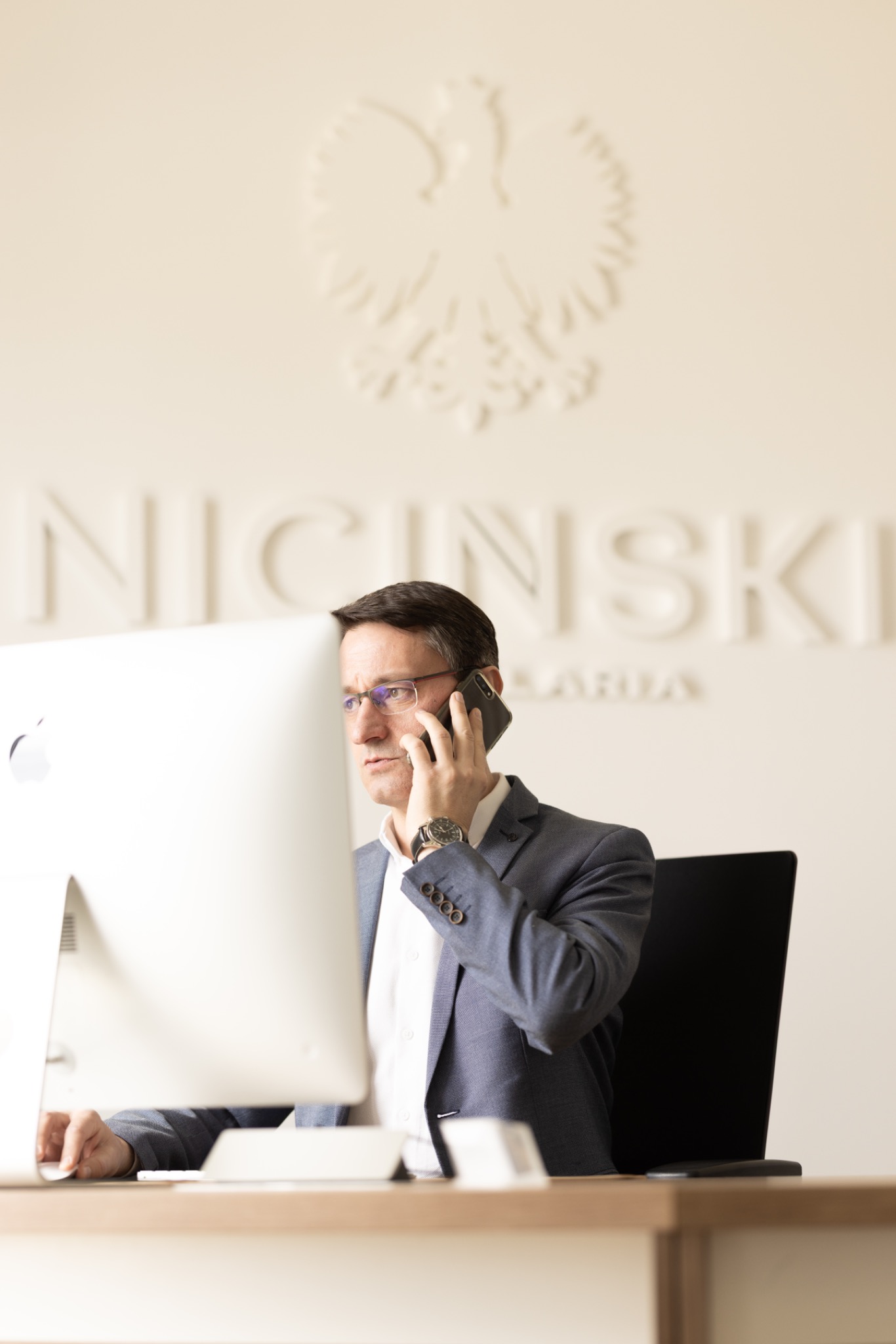 Prawnik w okularach rozmawia przez telefon w biurze w Łodzi. W tle godło Polski i nazwa kancelarii na ścianie, widoczny fragment komputera.