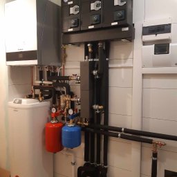 HYDROTOMS - Złożona instalacja grzewcza z piecem gazowym Buderus, zasobnikiem Elmell, rozdzielaczami Afriso i rurami w izolacji, zamontowana na tle białych płytek ściennych.