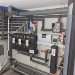 Rozbudowana instalacja grzewcza z licznymi rurami, zaworami, czujnikami i sterownikiem, widoczne logo QIK na kilku elementach, instalacja w pomieszczeniu technicznym.