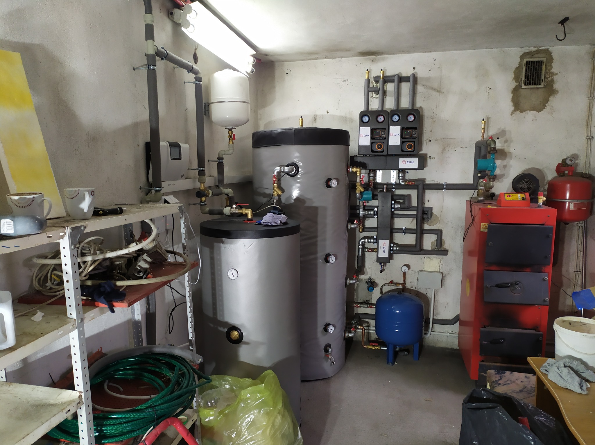 Zestaw urządzeń grzewczych w piwnicy: dwa zbiorniki akumulacyjne, piec CO na paliwo stałe, armatura, rury, manometry, zawory, oraz regał z narzędziami i wężami.