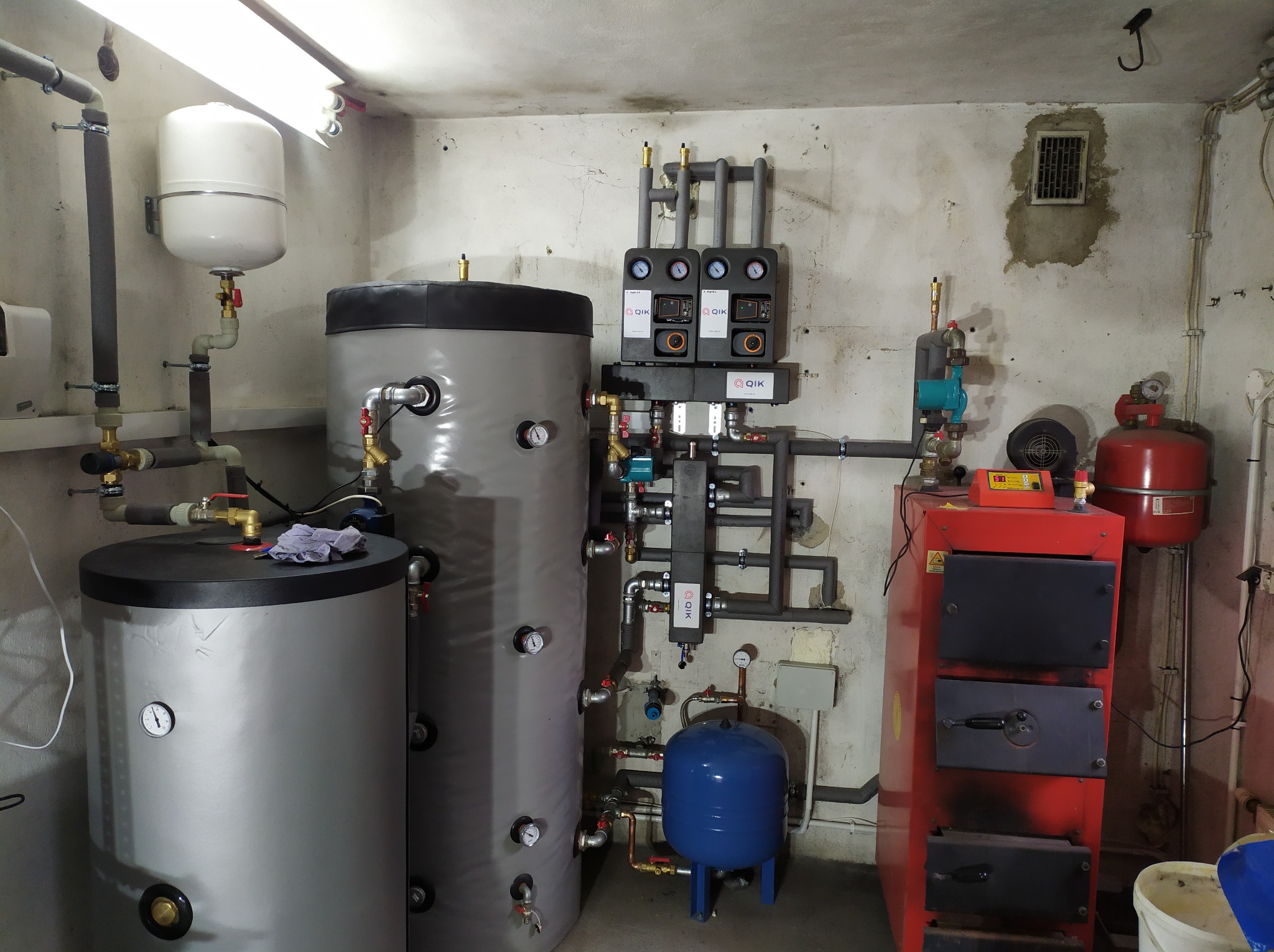 Kotłownia z zainstalowanym dwoma zbiornikami akumulacyjnymi, piecem na paliwo stałe i osprzętem hydraulicznym, na ścianie widoczne zawory i manometry.