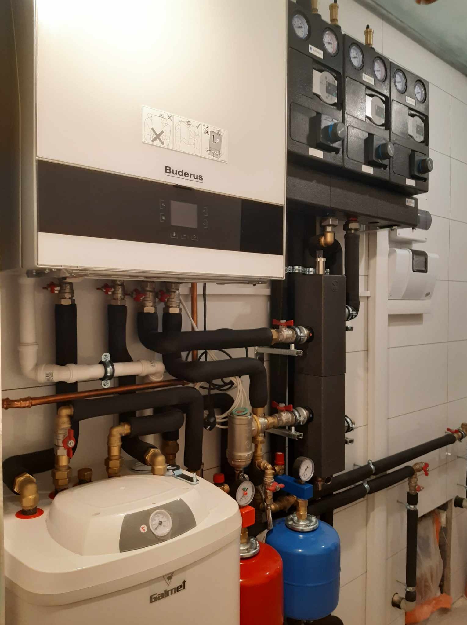 Biały piec gazowy Buderus z czarnym panelem, podłączony do instalacji z rur miedzianych i czarnych izolowanych, widoczne manometry i zawory, obok biały zbiornik Galmet i dwa zbiorniki ciśnieniowe...