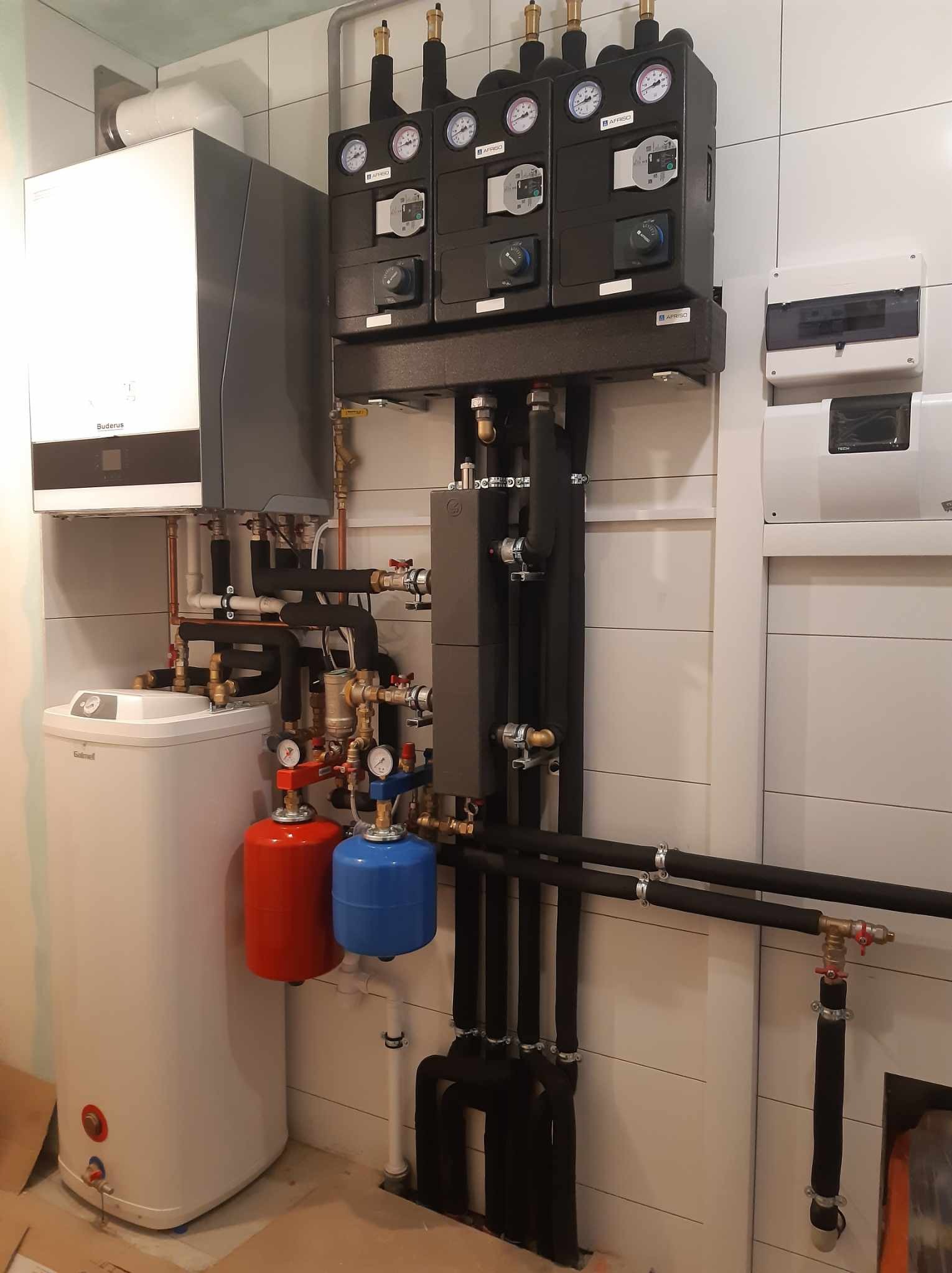 Złożona instalacja grzewcza z piecem gazowym Buderus, zasobnikiem Elmell, rozdzielaczami Afriso i rurami w izolacji, zamontowana na tle białych płytek ściennych.