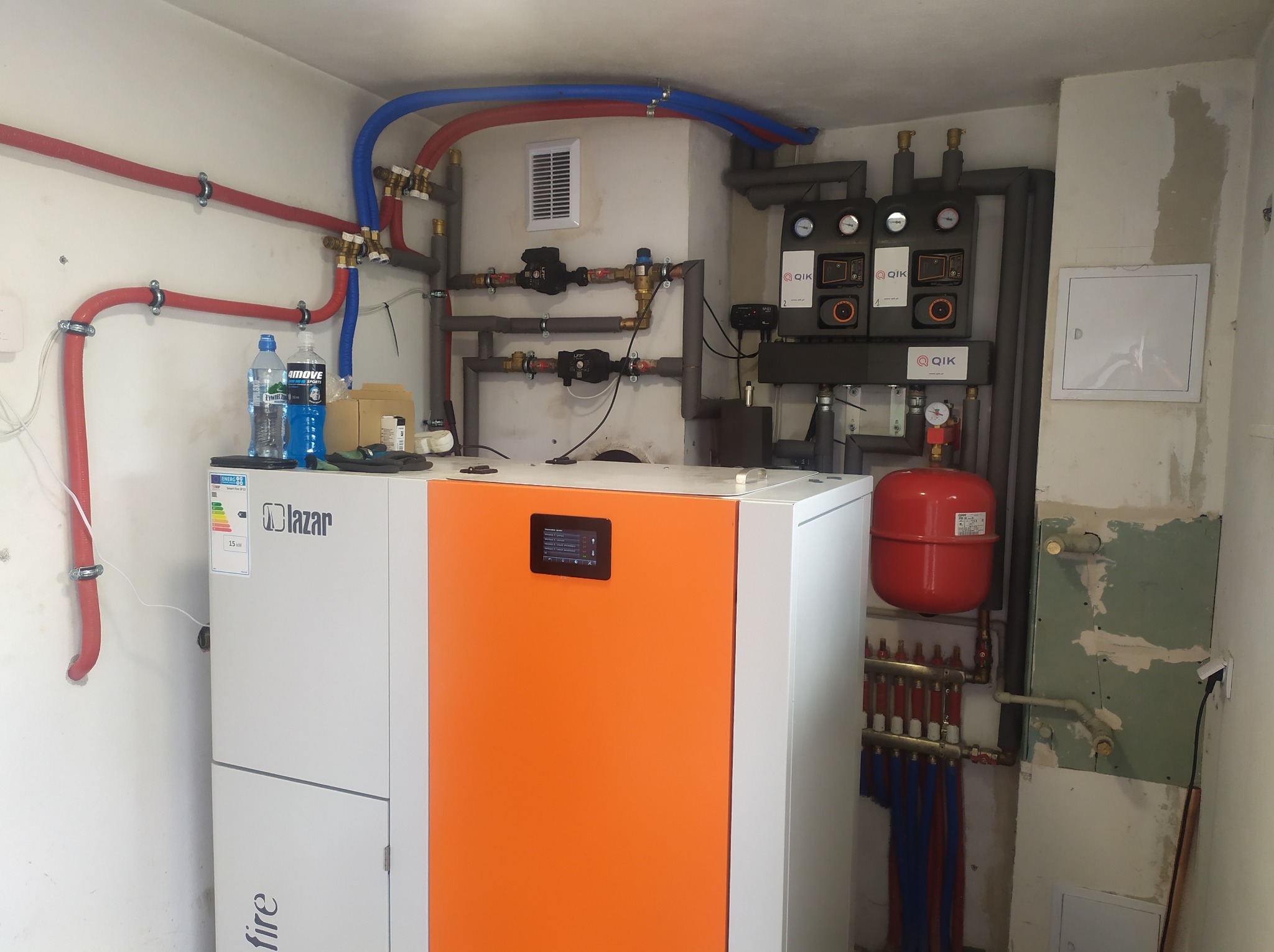 Nowoczesny piec gazowy Lazar z pomarańczowym panelem, instalacja grzewcza z rurami w kolorach czerwonym i niebieskim, rozdzielacz ogrzewania podłogowego, widoczne elementy sterujące QIK, butelki...