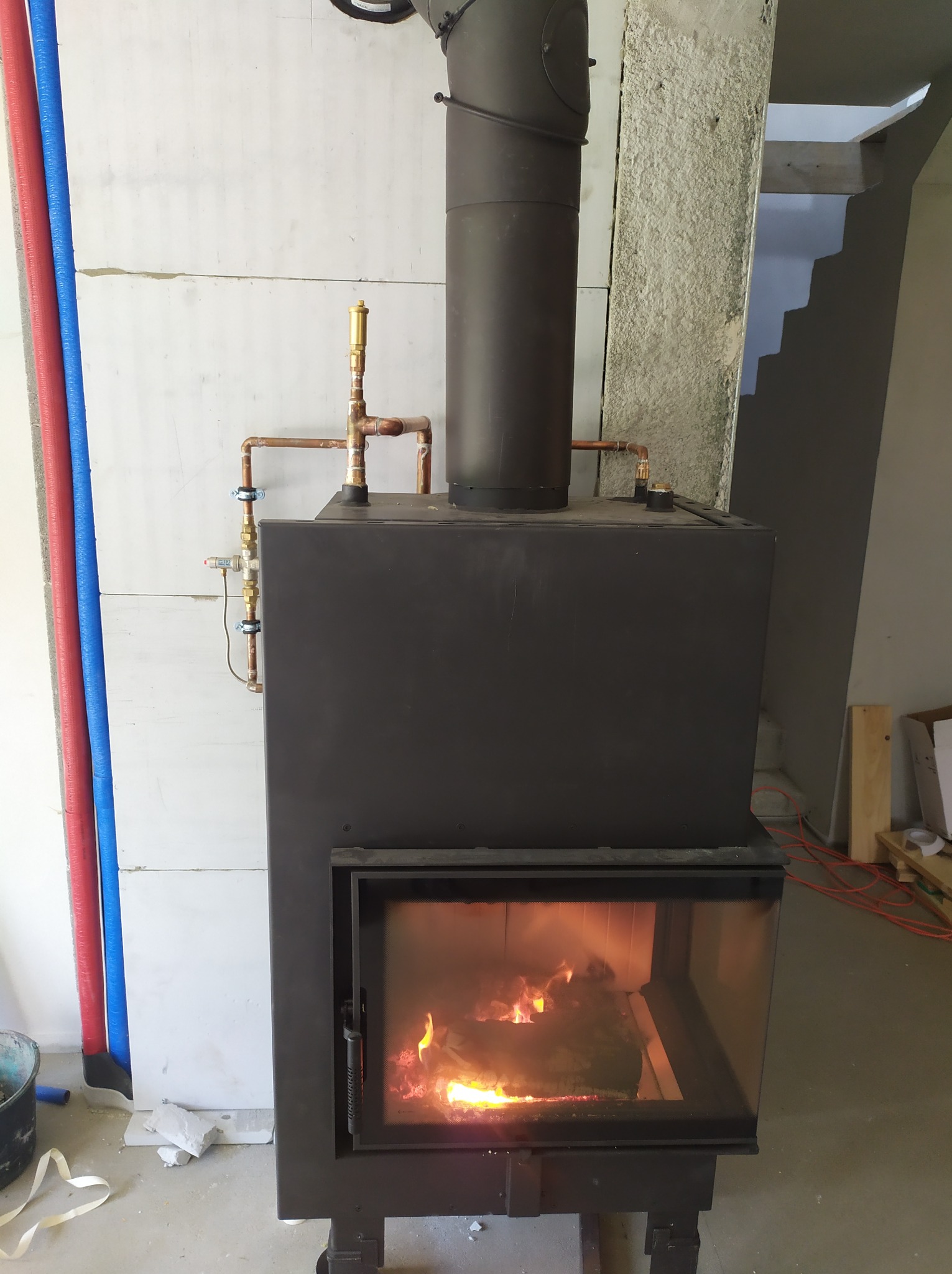 Czarny piec wolnostojący z palącym się drewnem wewnątrz, z widocznymi miedzianymi rurami i zaworami, na tle białych ścian z instalacją wodną w kolorze czerwonym i niebieskim, w surowym wnętrzu...