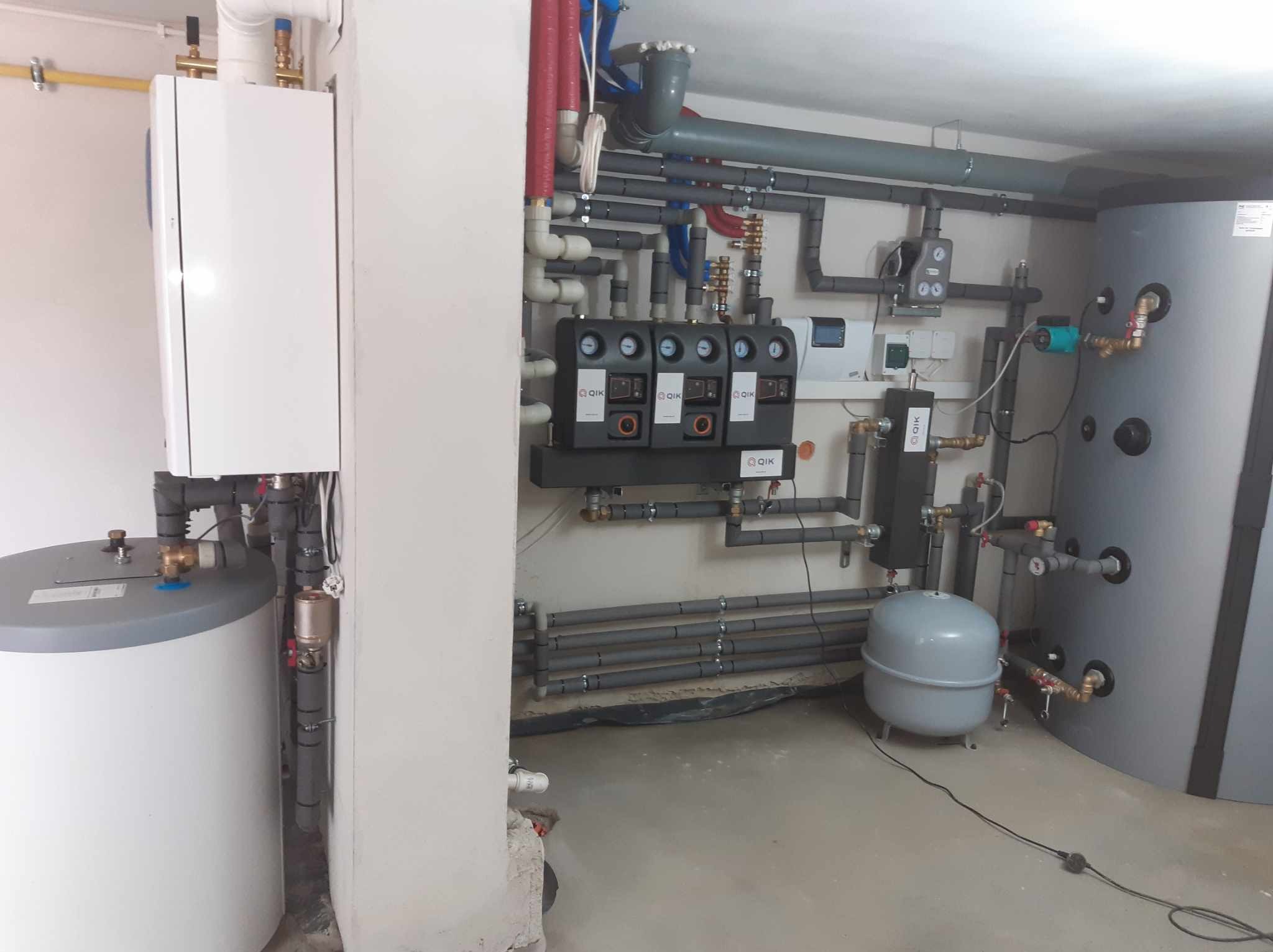Kotłownia z zainstalowanym piecem gazowym, zbiornikiem na wodę, zbiornikiem wyrównawczym i rozbudowaną instalacją hydrauliczną z licznymi rurami i zaworami.