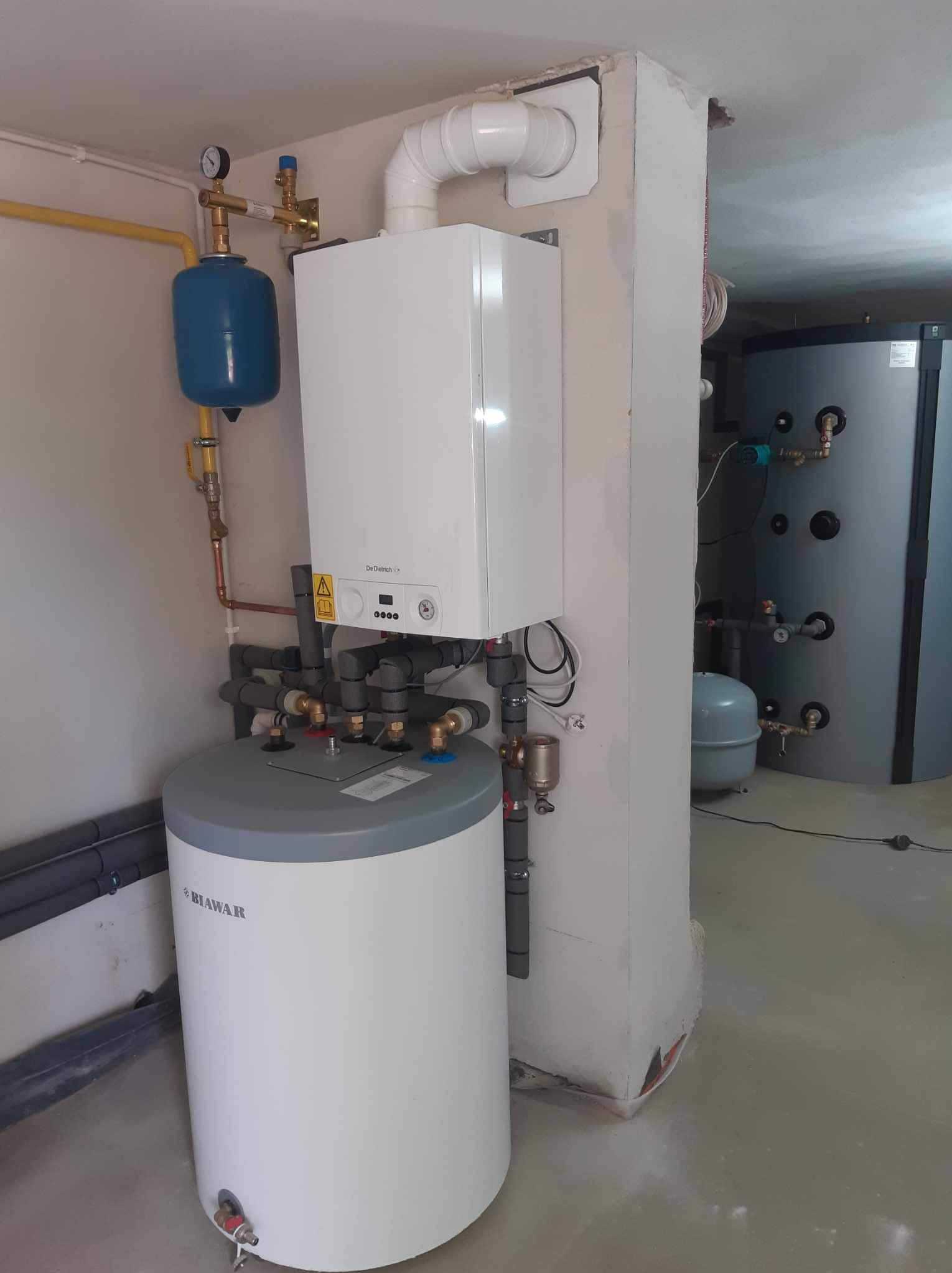 Biały piec gazowy De Dietrich z podłączonym zasobnikiem Biawar, niebieskie naczynie przeponowe, rury miedziane i szare rury PCV w kotłowni z betonową kolumną i szarym zbiornikiem w tle.