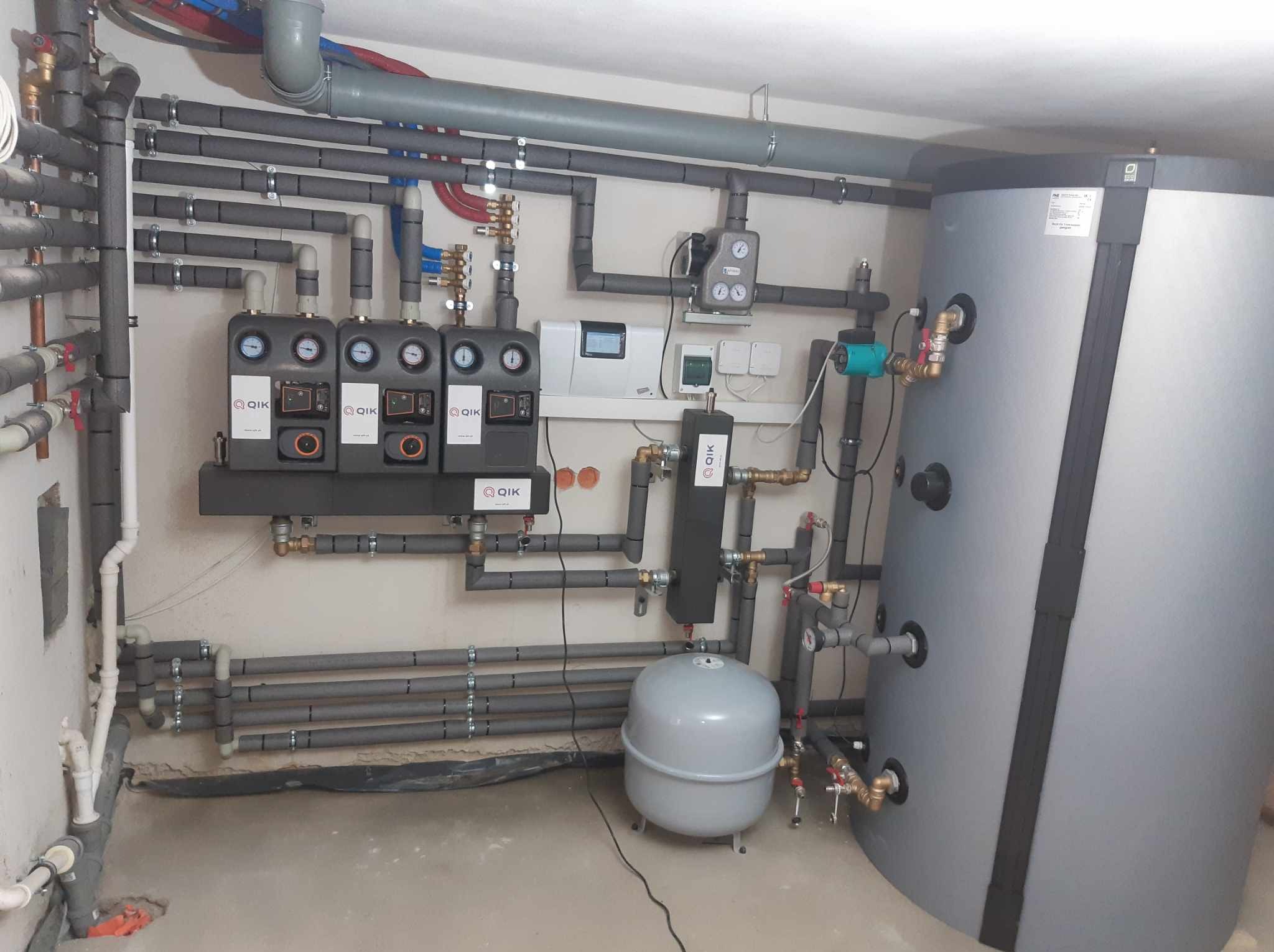 Złożona instalacja grzewcza w kotłowni, z szarymi rurami izolowanymi termicznie, zbiornikiem akumulacyjnym, oraz rozdzielaczami z manometrami, zamontowana na jasnej ścianie.