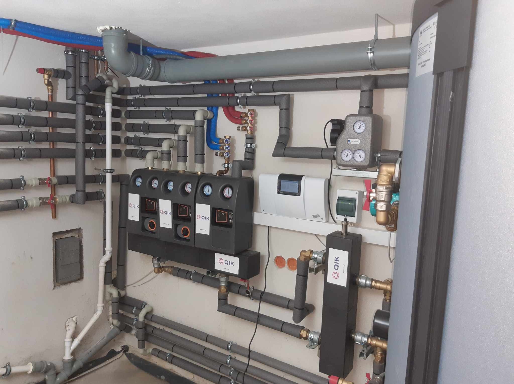 Rozbudowana instalacja grzewcza z licznymi rurami, zaworami, czujnikami i sterownikiem, widoczne logo QIK na kilku elementach, instalacja w pomieszczeniu technicznym.