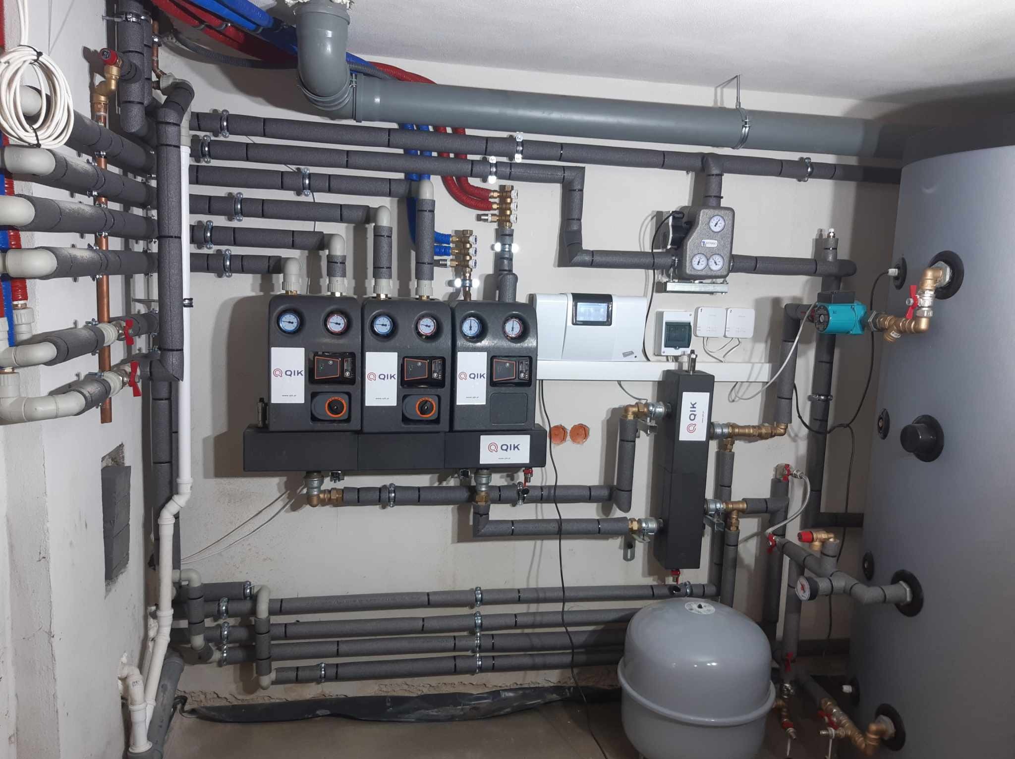 Złożona instalacja grzewcza w kotłowni z licznymi rurami, zaworami, pompami obiegowymi i zbiornikiem wyrównawczym, wszystko starannie zaizolowane i zamontowane na ścianie. Widoczny panel sterowania...