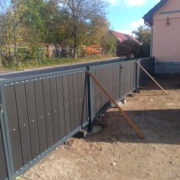 MJ - Fragment nowo montowanej bramy wjazdowej z ciemnymi panelami i szarą ramą, podpartej drewnianymi belkami, w tle widoczny dom i drzewa.