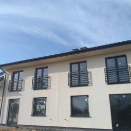 Stal Zachodniopomorskie - Nowoczesny dom dwukondygnacyjny z beżową elewacją, czarnymi oknami i balkonami zabezpieczonymi prostymi, stalowymi konstrukcjami, widok z poziomu gruntu na niezagospodarowany teren.