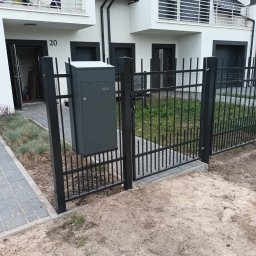 Stal Zachodniopomorskie - Nowoczesne ogrodzenie metalowe w kolorze grafitowym z bramką wejściową i skrzynką na listy, na tle budynku mieszkalnego z balkonem.