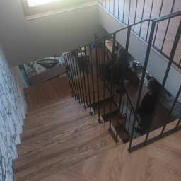 Stal Zachodniopomorskie - Widok z góry na drewniane schody z czarną metalową balustradą, wzorzysta tapeta na ścianie po lewej stronie, okno w tle.