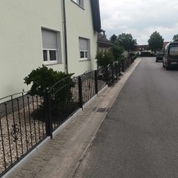 Stal Zachodniopomorskie - Czarny, metalowy płot z ozdobnymi elementami, oddzielający posesję od ulicy, widok na dom z roletami w oknach i zaparkowany samochód dostawczy w tle.