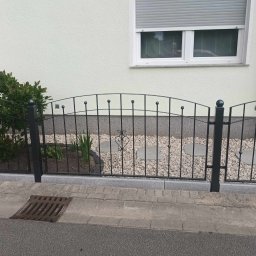 Stal Zachodniopomorskie - Czarne ogrodzenie z elementami dekoracyjnymi w kształcie serca, oddzielające ogródek z kamykami i roślinami od chodnika, na tle białej ściany budynku z oknem.