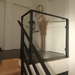 Stal Zachodniopomorskie - Szklana balustrada z czarnymi, metalowymi słupkami na schodach w jasnym wnętrzu. W tle widoczny dekoracyjny bukiet z suszonych traw i złoty zegar.