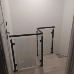 Szklana balustrada z czarnymi elementami mocującymi, zabezpieczona folią ochronną, zamontowana na schodach wewnątrz budynku, widok z góry.