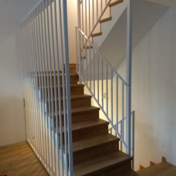 Stal Zachodniopomorskie - Drewniane schody z białą, pionową balustradą z metalowych prętów, widok z dołu, oświetlenie górne.