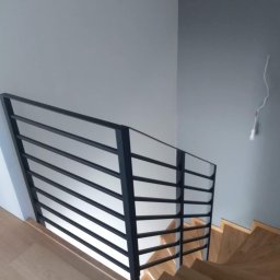 Stal Zachodniopomorskie - Minimalistyczna, czarna balustrada z poziomych prętów zamontowana przy drewnianych schodach w jasnym wnętrzu, widoczne niedokończone podłączenie elektryczne na ścianie.